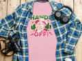 Garment Graphics presents a pink T-shirt with a bold Hands Off! floral motif and cowboy design, featuring a classic fit. Its elegantly displayed on wood beside a blue-green plaid shirt, headphones, white water bottle, and black camera.