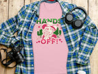 Garment Graphics presents a pink T-shirt with a bold Hands Off! floral motif and cowboy design, featuring a classic fit. Its elegantly displayed on wood beside a blue-green plaid shirt, headphones, white water bottle, and black camera.
