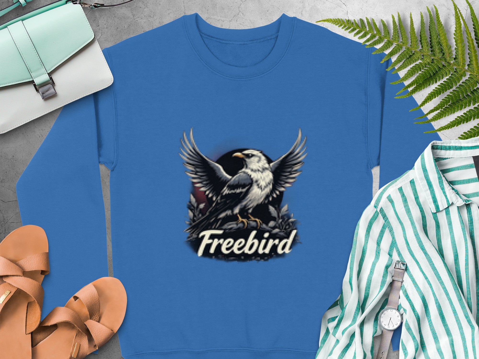 The blue unisex Freebird Graphic Sweatshirt by Garment Graphics displays an eagle with spread wings above the text. Its styled on a gray background with a green fern leaf, brown sandals, a purse, and a striped shirt accessorized with a watch.