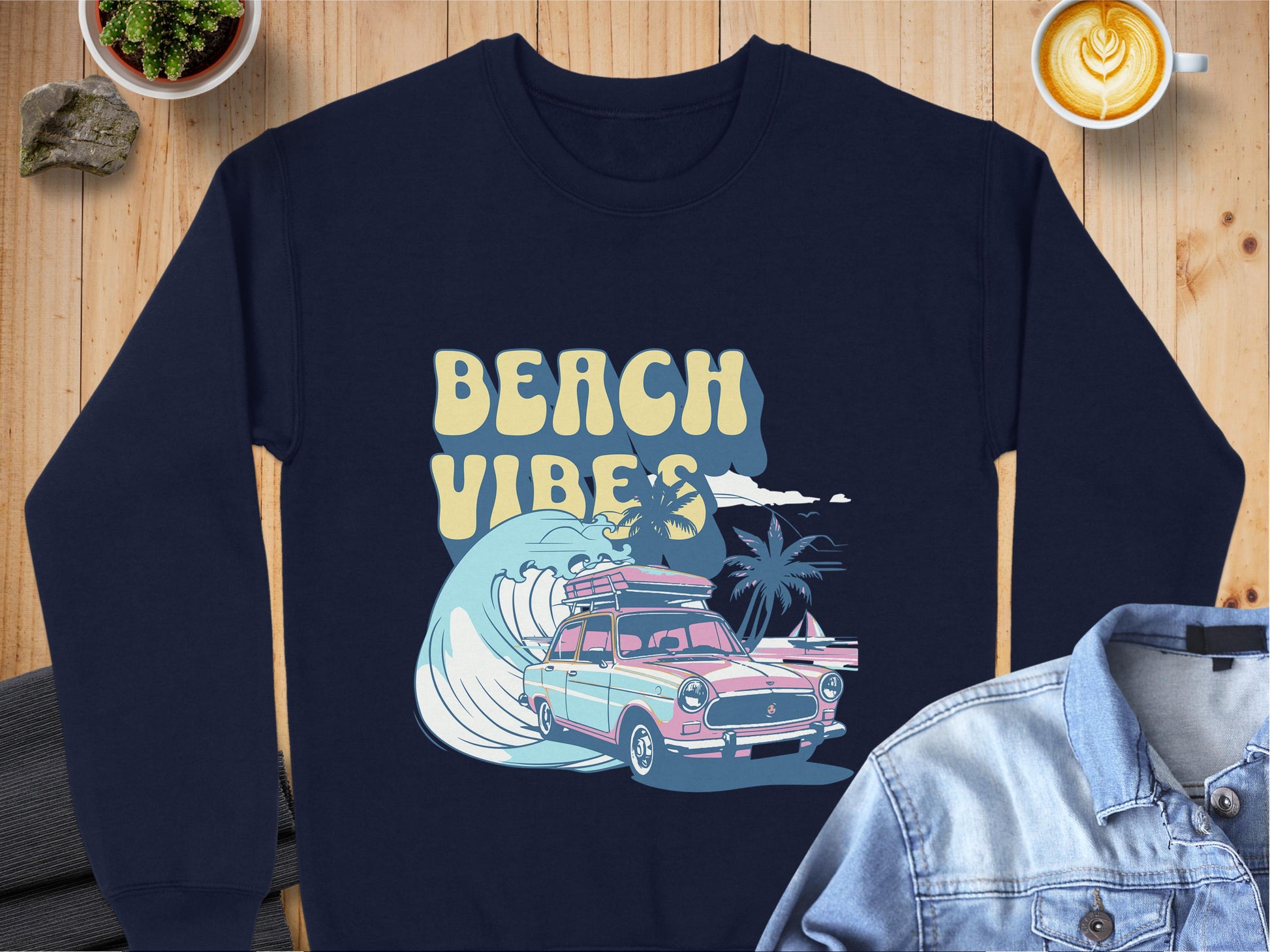 Garment Graphics navy sweatshirt boasts retro flair with its Beach Vibes design, featuring a classic car, surfboards, and palm trees on a wave. Its styled on wood with coffee, a denim jacket, and a small plant beside it.