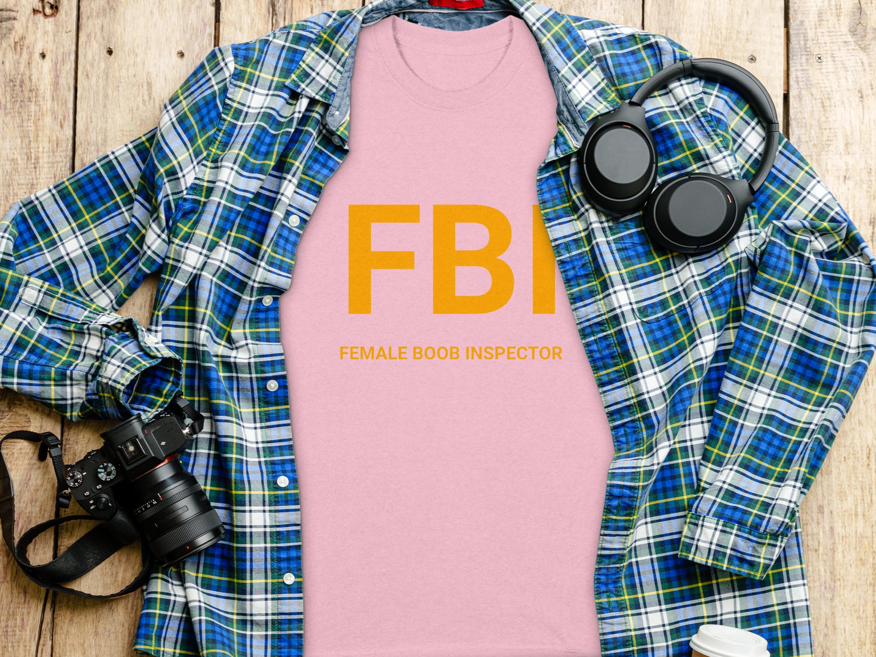 A flat lay photo features a Garment Graphics T-shirt with FBI Female Boob Inspector in yellow. The design is surrounded by a blue plaid shirt, headphones, a camera, and a cup on a wooden surface, highlighting the humorous style.