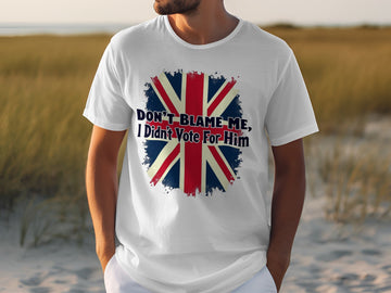 Wearing a Garment Graphics classic fit white T-shirt featuring the Union Jack and the text Dont blame me, I didnt vote for him, someone makes a bold statement. The backdrop features sandy terrain with grass patches.