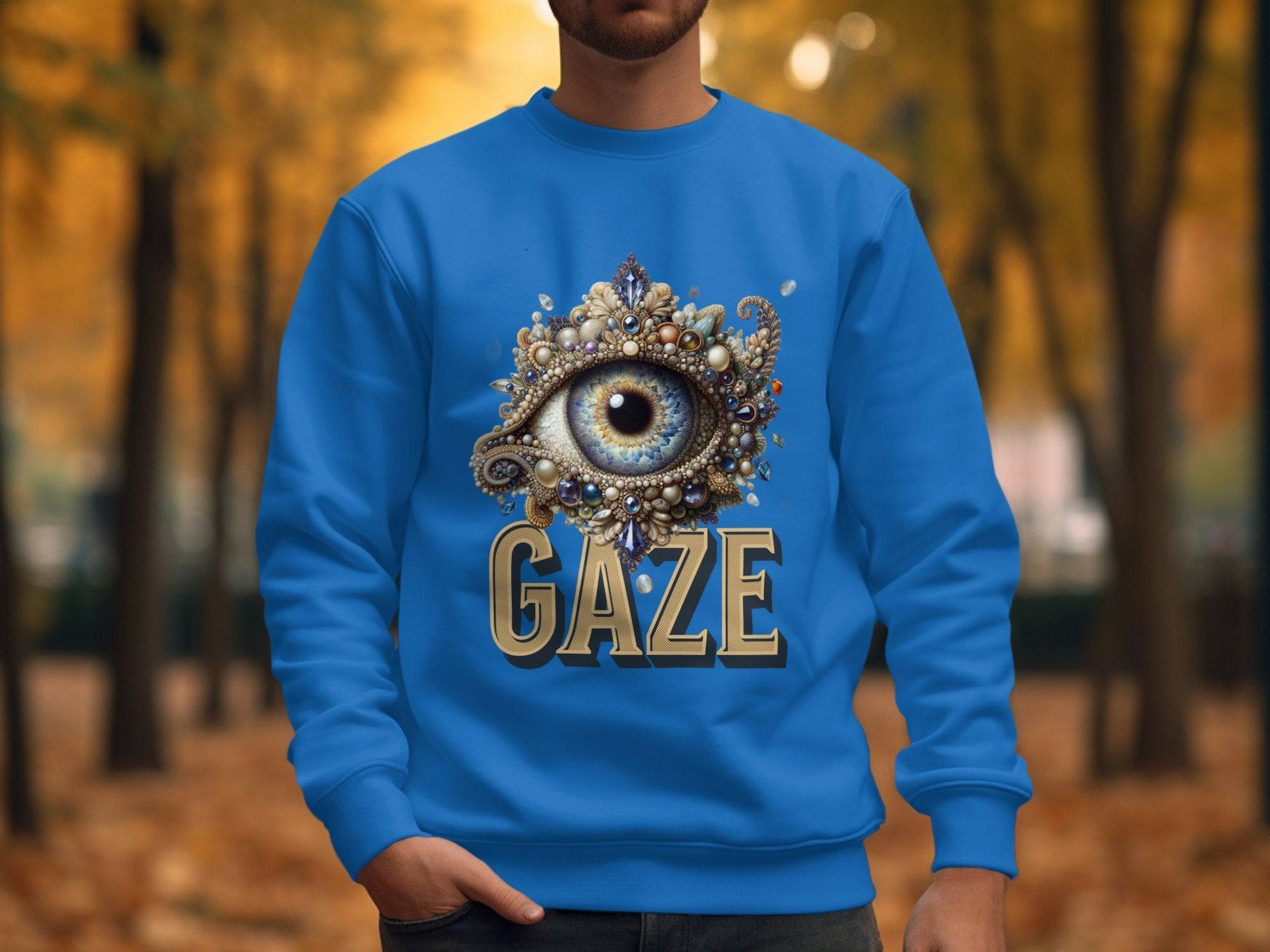 A person in a bright blue Garment Graphics sweatshirt with an ornate eye design above GAZE stands on a tree-lined path covered in autumn leaves. The unisex sizing ensures comfort amid the warm, golden backdrop.