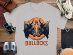 A light gray T-shirt by Garment Graphics, from the Bullocks Pintable T-Shirts collection, showcases two bulls facing each other on an orange circle with BULLOCKS below. The classic fit shirt is displayed on a wooden surface amidst assorted items.