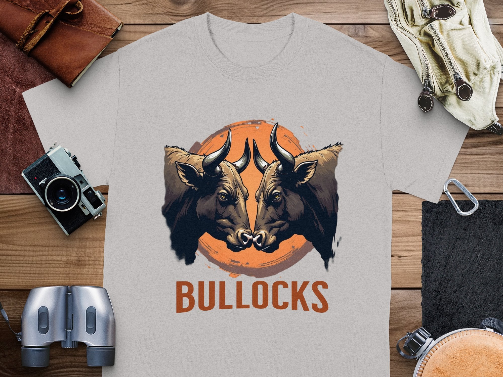 A light gray T-shirt by Garment Graphics, from the Bullocks Pintable T-Shirts collection, showcases two bulls facing each other on an orange circle with BULLOCKS below. The classic fit shirt is displayed on a wooden surface amidst assorted items.
