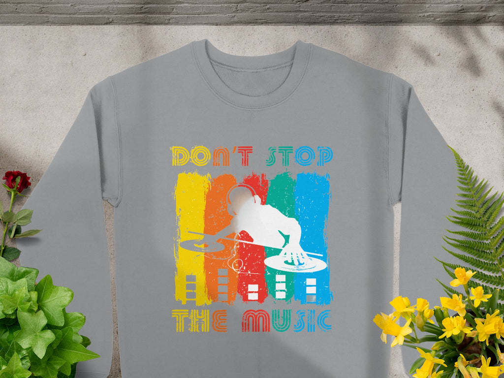 Gray sweatshirt by Garment Graphics with a vibrant DJ design over vertical yellow, red, green, and blue stripes. Features retro Dont Stop the Music text above and below the colorful DJ graphic using turntables. Surrounded by plants, it’s perfect for music lovers.