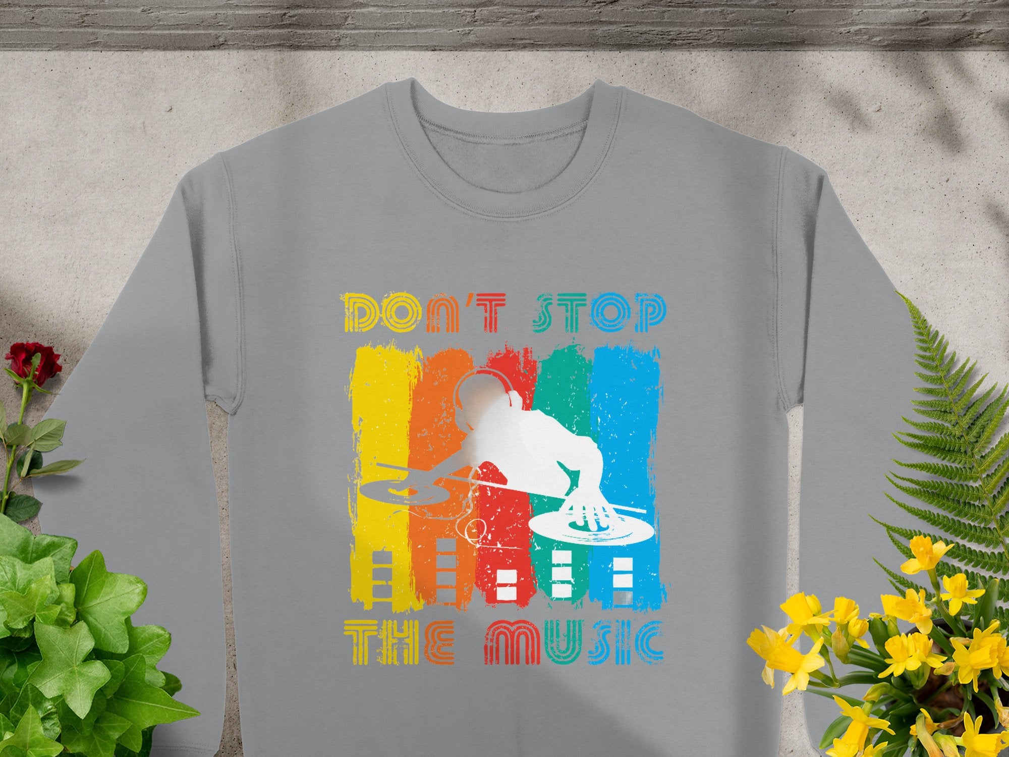 Gray sweatshirt by Garment Graphics with a vibrant DJ design over vertical yellow, red, green, and blue stripes. Features retro Dont Stop the Music text above and below the colorful DJ graphic using turntables. Surrounded by plants, it’s perfect for music lovers.