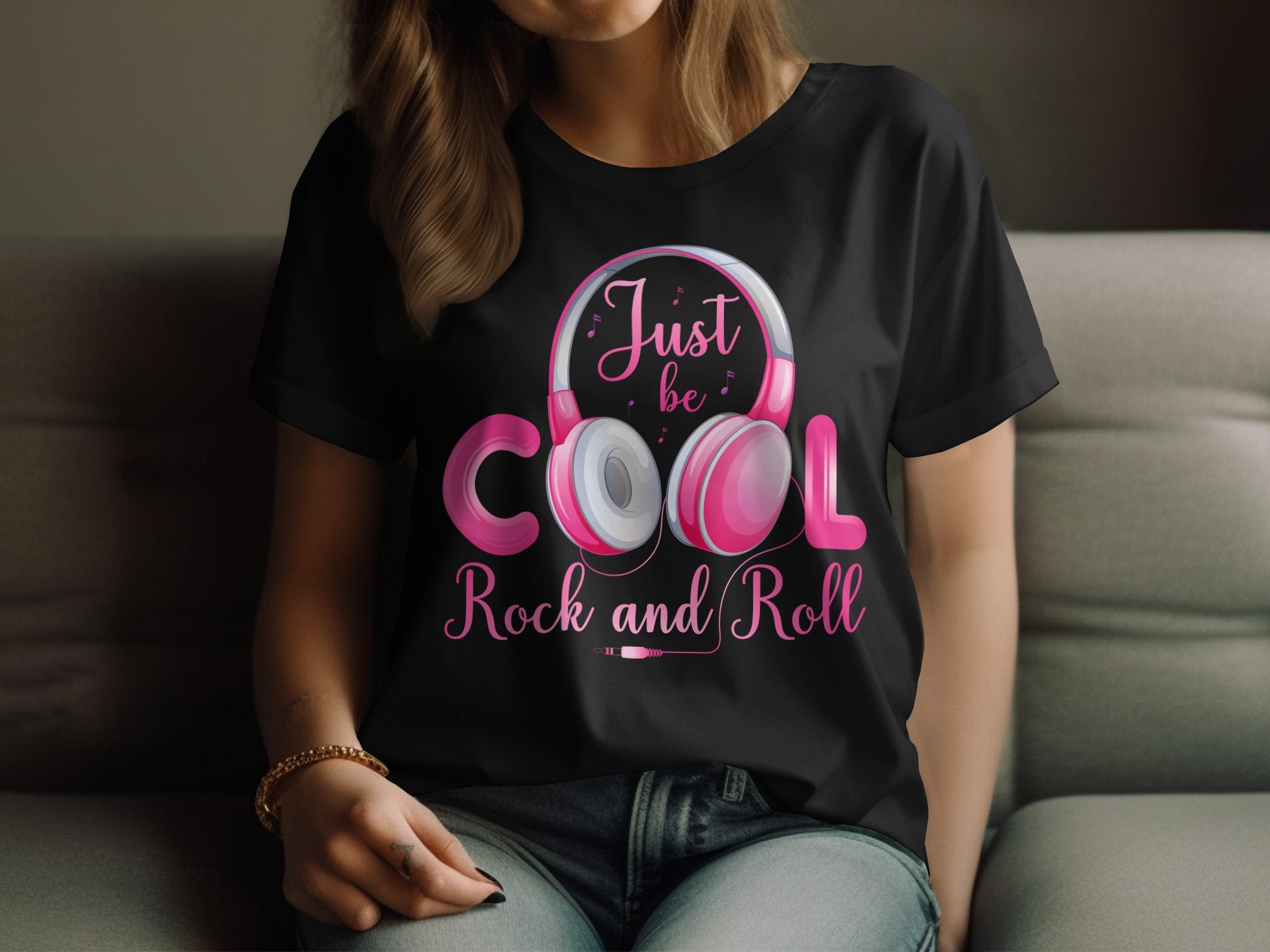 A person with brown hair sits on a couch, wearing a Garment Graphics black T-shirt featuring pink headphones and the text Just be Cool Rock and Roll in pink and white. Their hand rests on their knee, exuding relaxed charm, making this tee ideal for music lovers.