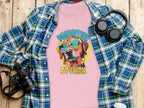 Garment Graphics pink t-shirt showcases a vibrant dog-in-sunglasses graphic and Pawsitive Stylish text. Perfect for dog lovers, its displayed on wood with a blue plaid shirt, headphones, camera, and coffee cup. Womens sizing.