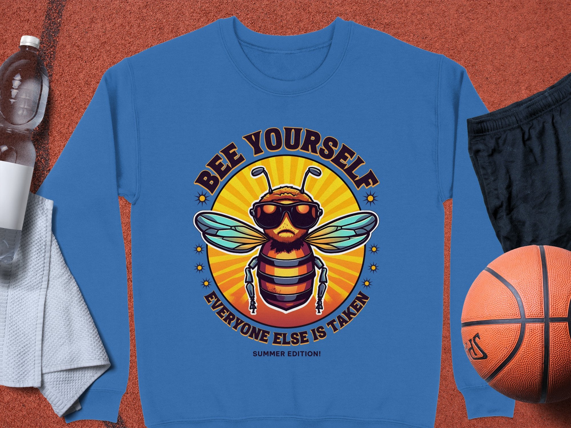 The Garment Graphics Summer Edition sweatshirt displays a fun bee in sunglasses with the phrase Bee Yourself, Everyone Else is Taken! Great for all with unisex sizing, its pictured alongside a water bottle, white towel, black shorts, and a basketball.