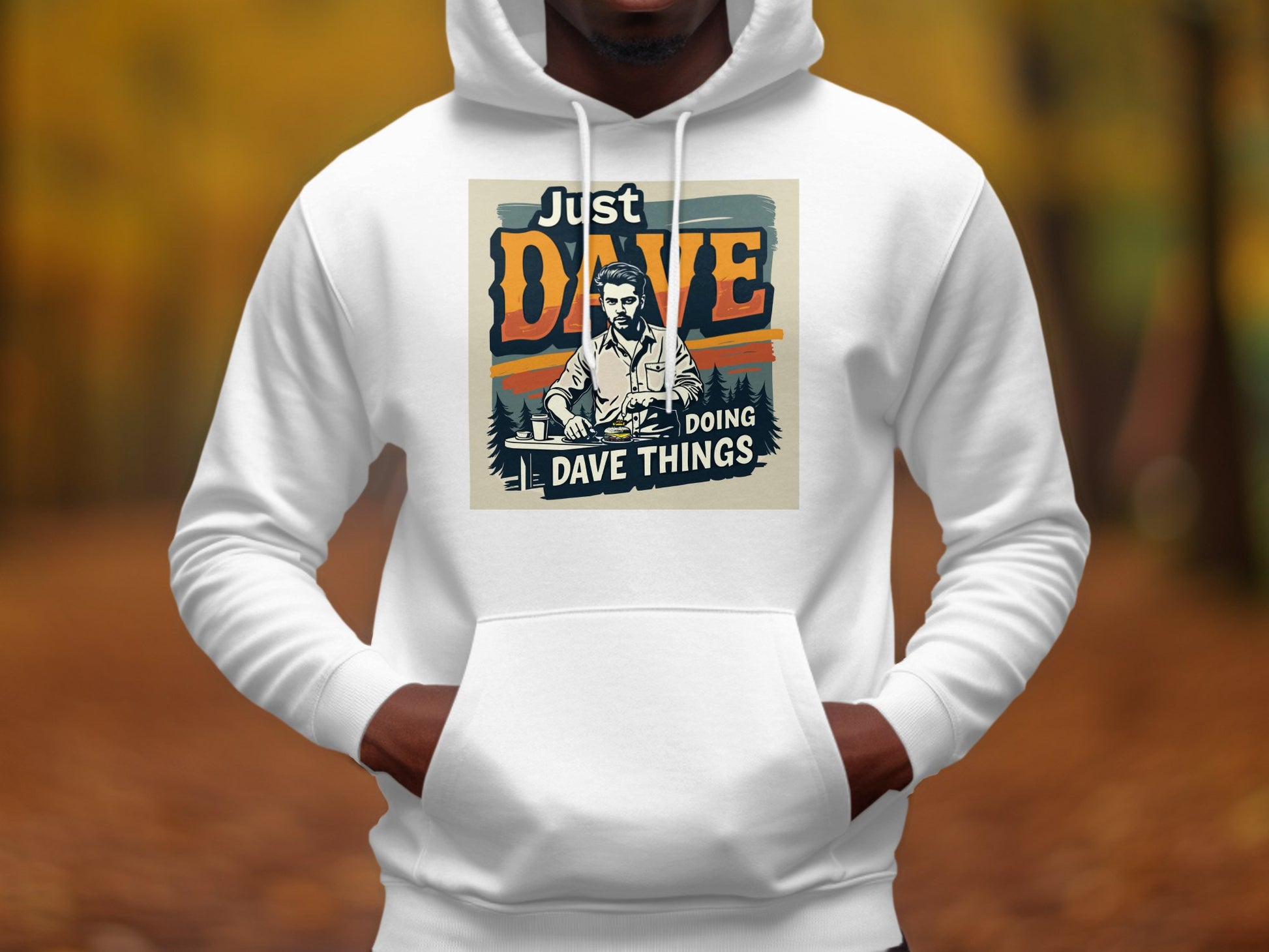 A person wears a white medium-heavy fabric graphic hoodie by Garment Graphics, featuring an illustration of a man with the phrase Just Dave Doing Dave Things, set against a blurred outdoor scene with autumn-colored leaves.