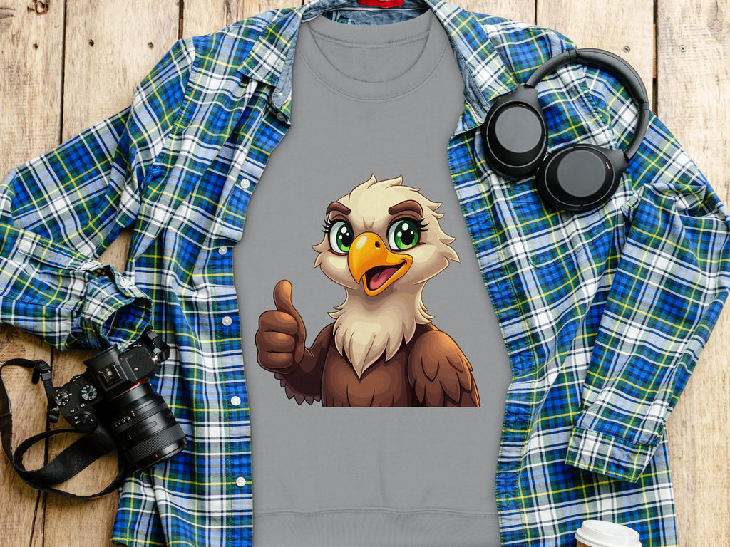 A Garment Graphics gray shirt with a cartoon eagle giving a thumbs up layers over a blue plaid shirt. The unisex fit suits everyone. Nearby, theres a black camera, headphones, and a to-go coffee cup on a wooden surface.