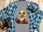 A Garment Graphics gray shirt with a cartoon eagle giving a thumbs up layers over a blue plaid shirt. The unisex fit suits everyone. Nearby, theres a black camera, headphones, and a to-go coffee cup on a wooden surface.