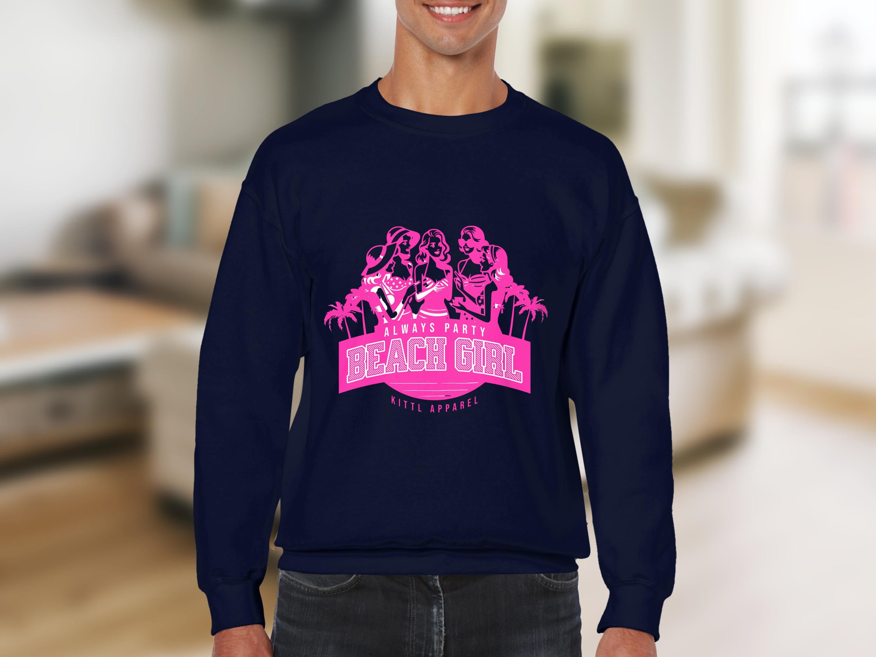 A person is wearing a dark blue sweatshirt from Garment Graphics, featuring a pink graphic of three cartoon women dancing among palm trees. The design includes the text Always Party BEACH GIRL. The background is softly blurred.
