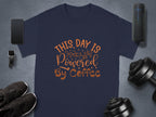 A flat lay photo features a Garment Graphics classic fit navy t-shirt with This Day is Powered by Coffee in brown. A smartphone, fitness tracker, wireless speaker, shoes, and dumbbell weights surround the coffee t-shirt on a gray background.
