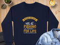 A navy blue Garment Graphics sweatshirt features Father And Son Fishing Partners For Life in yellow and white with a fishing boat and fish graphic. It rests on a wooden surface surrounded by a stone, plant, latte, and denim jacket.