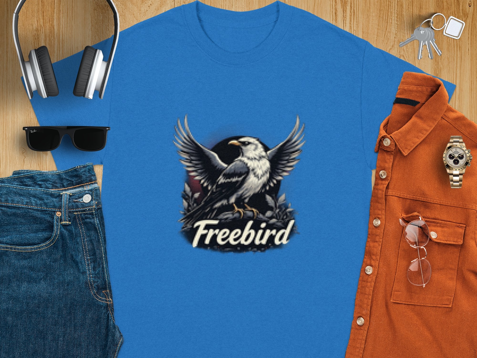 A flat lay image showcases a Garment Graphics eagle T-shirt in blue, crafted from 100% cotton with a bold graphic design. Accompanying items include headphones, sunglasses, jeans, a brown shirt, a watch, and keys scattered on the wooden surface.
