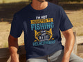 A man sits on a wooden bench wearing a Garment Graphics T-shirt, featuring the fun text: Im not addicted to fishing; we are just in a very committed relationship in various fonts and colors. The blurred outdoor backdrop suits dedicated anglers perfectly.