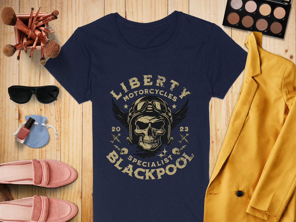 A Garment Graphics navy blue T-shirt with a skull, wings design, and Liberty Motorcycles Specialist Blackpool 2023 text is on wood. Nearby are a yellow jacket, makeup brushes, eyeshadow palette, sunglasses, pink shoes, and nail polish.