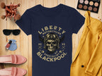 A Garment Graphics navy blue T-shirt with a skull, wings design, and Liberty Motorcycles Specialist Blackpool 2023 text is on wood. Nearby are a yellow jacket, makeup brushes, eyeshadow palette, sunglasses, pink shoes, and nail polish.