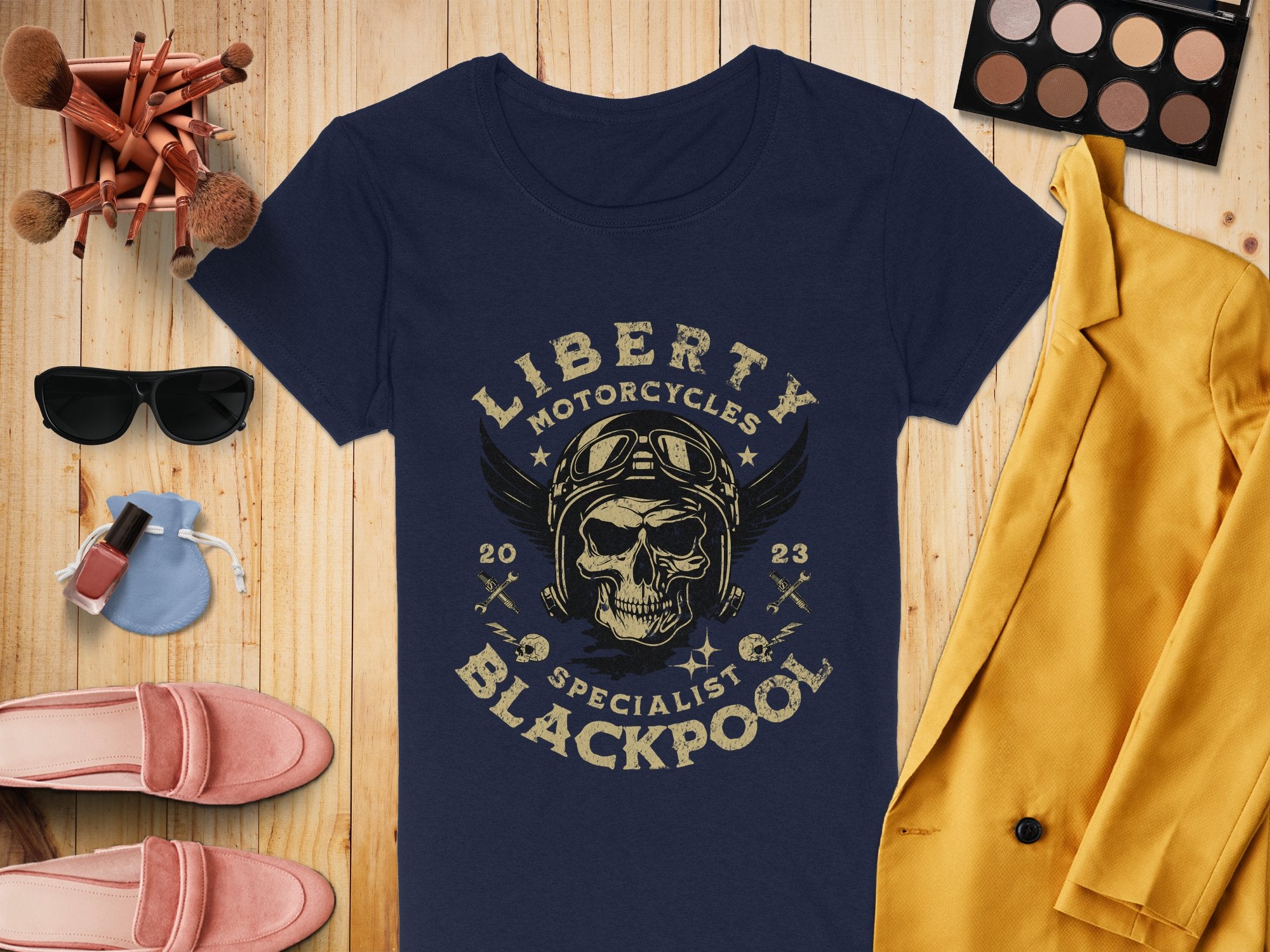 A Garment Graphics navy blue T-shirt with a skull, wings design, and Liberty Motorcycles Specialist Blackpool 2023 text is on wood. Nearby are a yellow jacket, makeup brushes, eyeshadow palette, sunglasses, pink shoes, and nail polish.