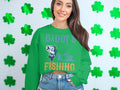 A woman wearing Garment Graphics jeans and a green Daddys Fishing Buddy sweater adorned with playful fish and boat illustrations stands against a wall of green shamrocks, her long brown hair adding charm to this perfect outfit for fishing enthusiasts.