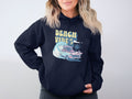 A person wears a Garment Graphics navy hoodie featuring a vintage car and surfboard graphic on a beach, with palm trees, waves, and Beach Vibes text. They have shoulder-length blonde hair and stand against a plain background.