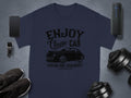 Garment Graphics presents a dark blue T-shirt with the Enjoy Classic Car slogan and a vintage car image. Ideal for car lovers, it sits on a gray surface with a smartphone, smartwatch, speaker, black shoes, and dumbbells.