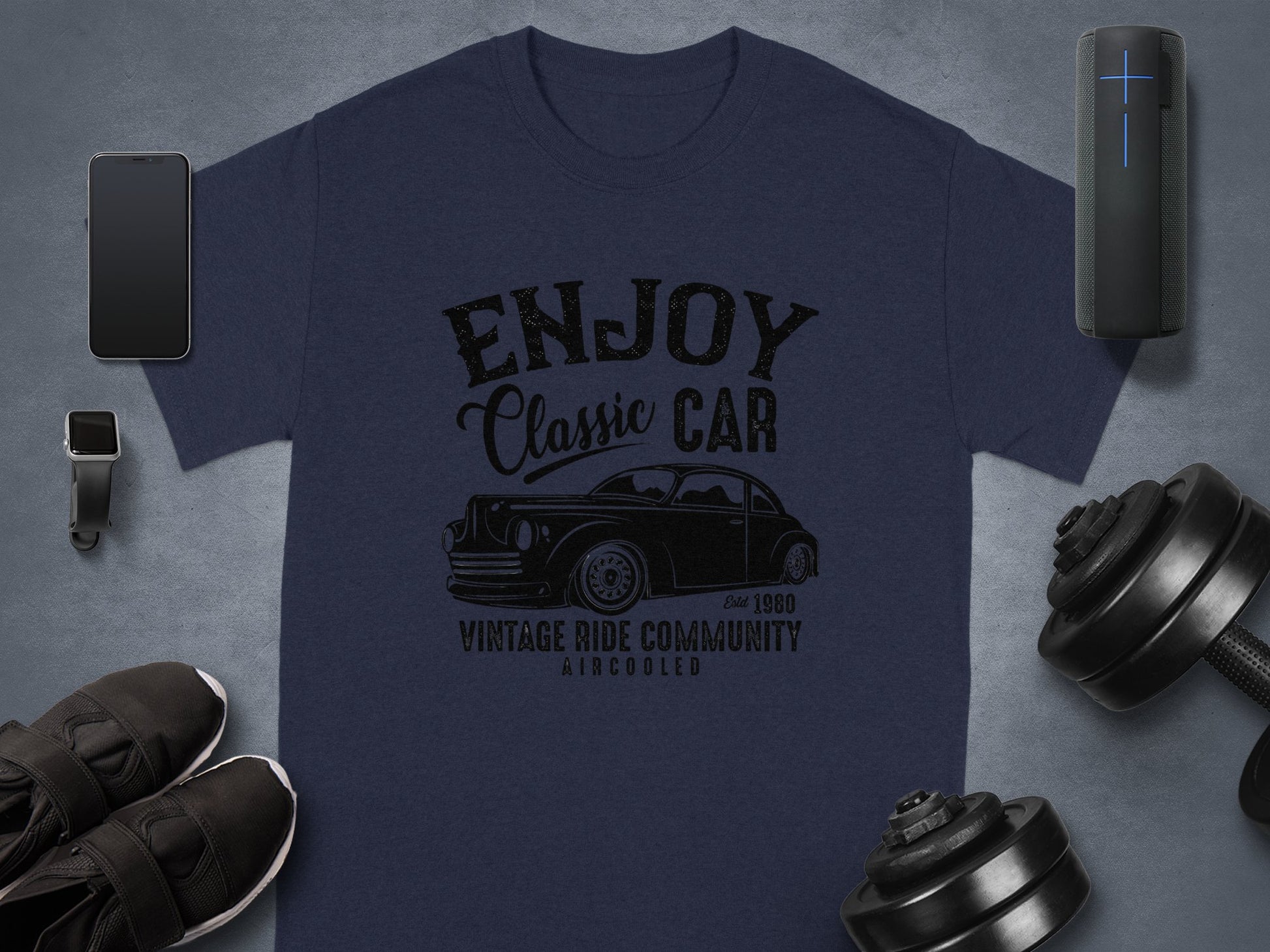 Garment Graphics presents a dark blue T-shirt with the Enjoy Classic Car slogan and a vintage car image. Ideal for car lovers, it sits on a gray surface with a smartphone, smartwatch, speaker, black shoes, and dumbbells.