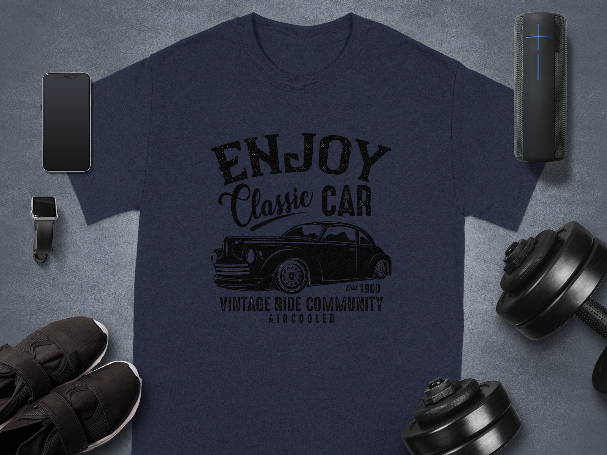 Garment Graphics presents a dark blue T-shirt with the Enjoy Classic Car slogan and a vintage car image. Ideal for car lovers, it sits on a gray surface with a smartphone, smartwatch, speaker, black shoes, and dumbbells.