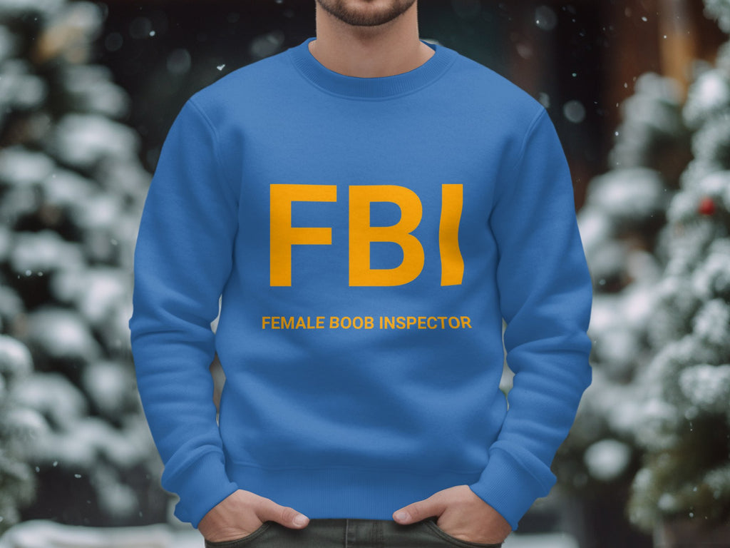 A person is wearing a blue Garment Graphics sweatshirt featuring FBI in bold yellow letters and the playful phrase FEMALE BOOB INSPECTOR. This unisex design suits anyone. Snow-covered trees are visible in the blurred background, enhancing the lighthearted scene.