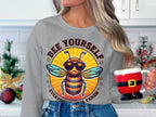 In a kitchen, someone with long hair holds a Santa-themed mug while wearing Garment Graphics summer edition gray sweatshirt featuring a bee in sunglasses and the quote, Bee Yourself, Everyone Else Is Taken, available in unisex sizes.