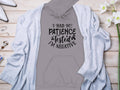 The Garment Graphics Patience Tested Hoodie, made from medium-heavy fabric in a classic fit, features the text I had my patience tested, Im negative. It pairs well with a light grey cardigan, white high heels, jewelry and watch, accented by white flowers on a wooden surface.