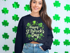 A person wearing Garment Graphics black shamrock graphic sweatshirt with a Happy St. Patricks Day design, featuring a leprechaun hat and cheerful text, stands in front of green shamrocks. Smiling with long hair, they radiate festive cheer.