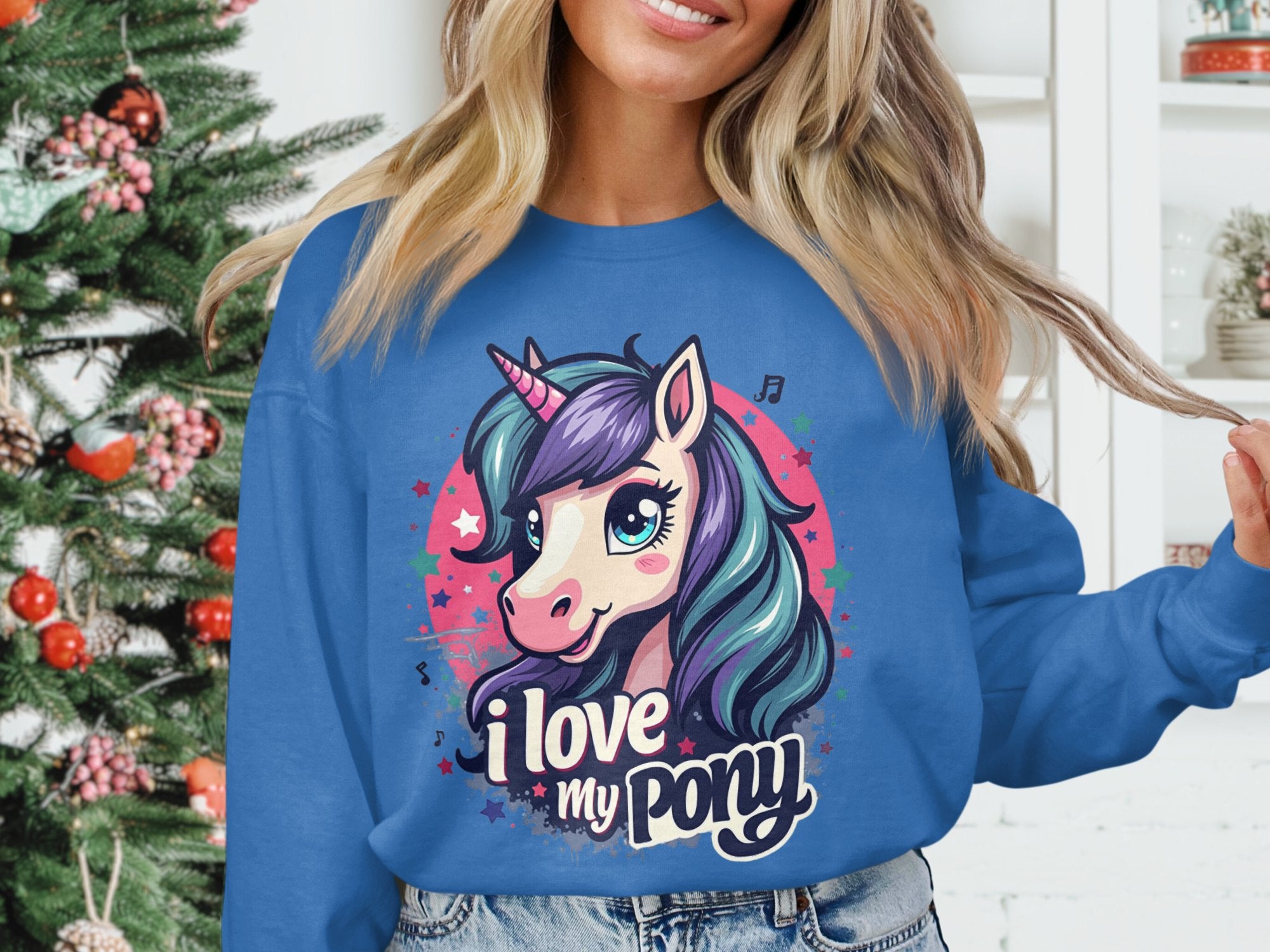 A person wears a vibrant Garment Graphics sweatshirt showcasing a colorful pony-unicorn with the phrase I love my pony. A decorated Christmas tree is partially visible in the background.