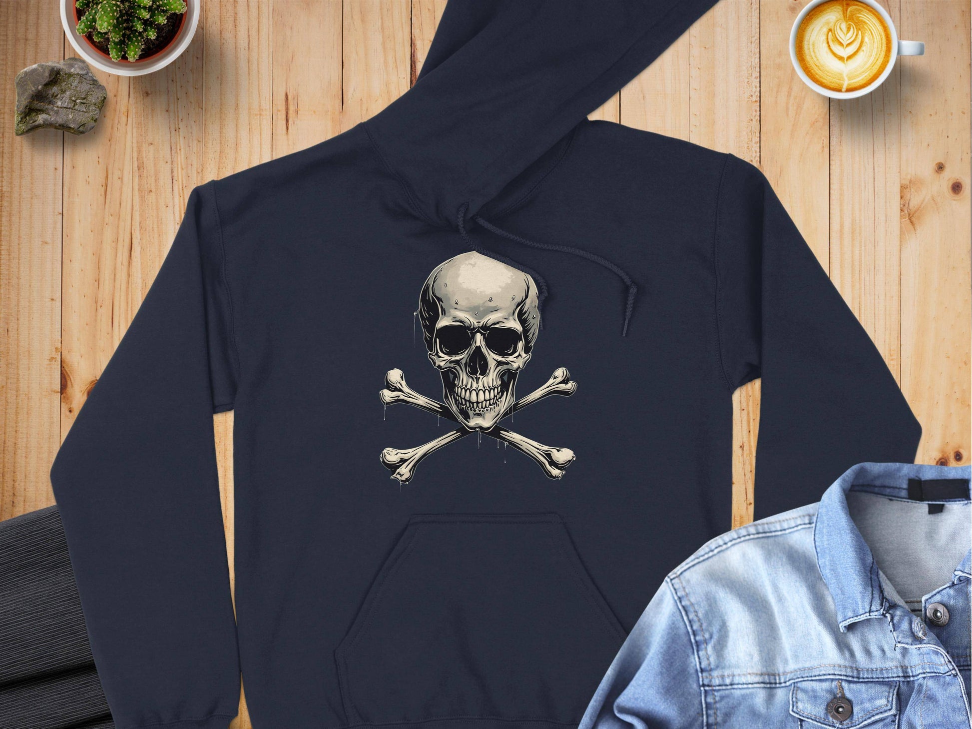 A Garment Graphics black hoodie with a skull and crossbones design rests on a wooden surface, surrounded by gray pants, a denim jacket, a latte with heart foam, and a small potted plant.