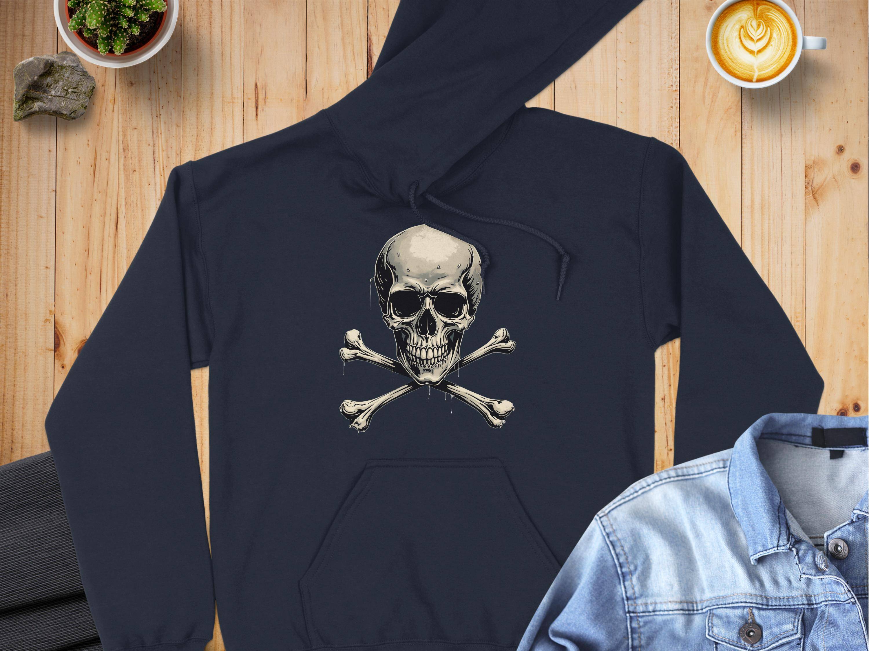 A Garment Graphics black hoodie with a skull and crossbones design rests on a wooden surface, surrounded by gray pants, a denim jacket, a latte with heart foam, and a small potted plant.