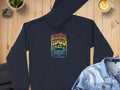 A Garment Graphics retro black hoodie featuring a vibrant Woodstock 1969 graphic with a peace sign and flowers is displayed on wood. Accompanied by a blue denim jacket, coffee latte art, and a small plant, it creates the perfect scene.