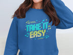 A person wears a Garment Graphics unisex, comfortable sweatshirt featuring the phrase Take it Easy in colorful letters with musical notes and arrows. The persons face is not visible.