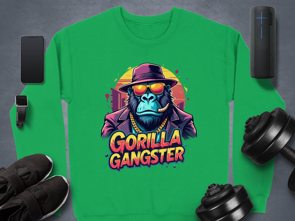 Green Garment Graphics urban sweatshirt with a gorilla graphic sporting sunglasses and a hat, labeled Gorilla Gangster. Nearby are a smartphone, smartwatch, wireless speaker, sneakers, and dumbbells on a dark surface.