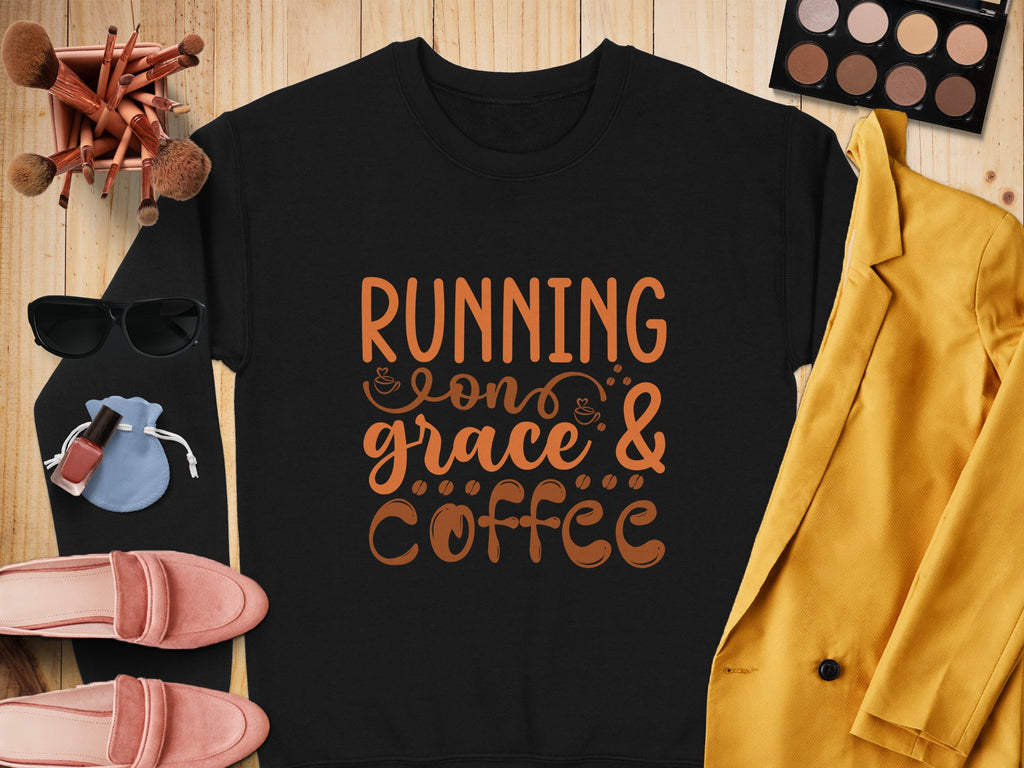 A flat lay features a Garment Graphics black graphic sweatshirt reading Running on grace & coffee in unisex sizing, surrounded by pink shoes, yellow jacket, sunglasses, makeup brushes, eyeshadow palette, lipsticks, and a small blue pouch on a wooden surface.