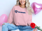 A person in a Garment Graphics pink SCANIA logo sweatshirt sits cross-legged, ideal for casual outings. Holding a red rose, theyre surrounded by two pink heart-shaped balloons. Their long wavy hair and ripped blue jeans highlight the charm of unisex sizing and effortless style.