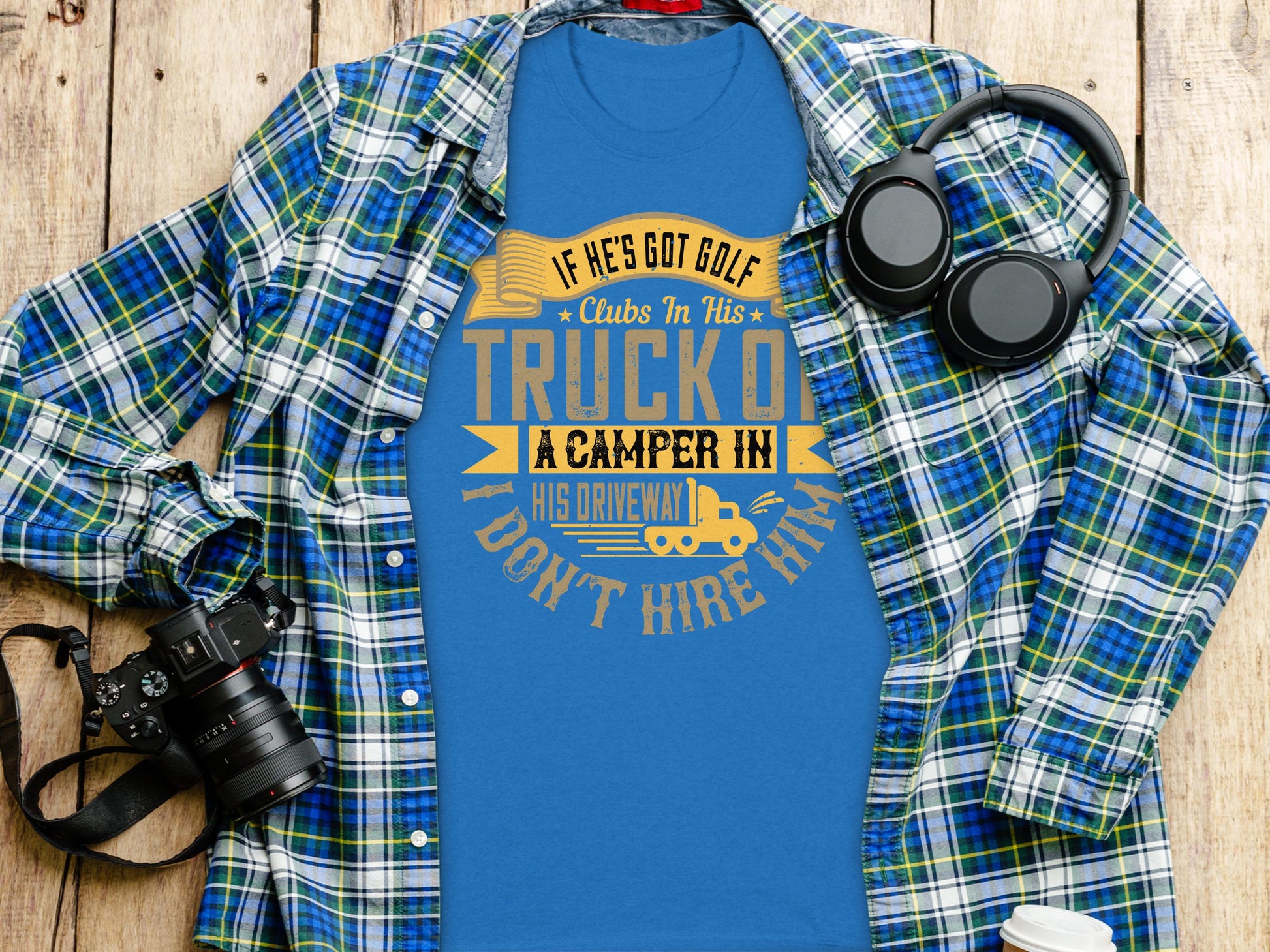 Flat lay of a Garment Graphics blue tee with a yellow humorous quote under a blue and green plaid shirt, alongside black headphones and a camera on wood. Ideal gift for style-savvy humor lovers.