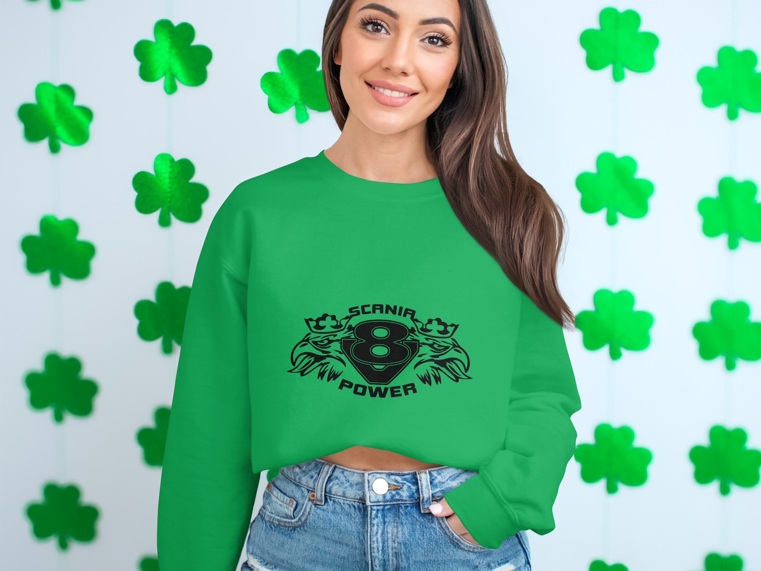 A stylish woman with long brown hair wears a green Garment Graphics Scania V8 Power sweatshirt featuring a cool design. She stands in front of a white wall decorated with green shamrock cutouts, completing her look with denim shorts.