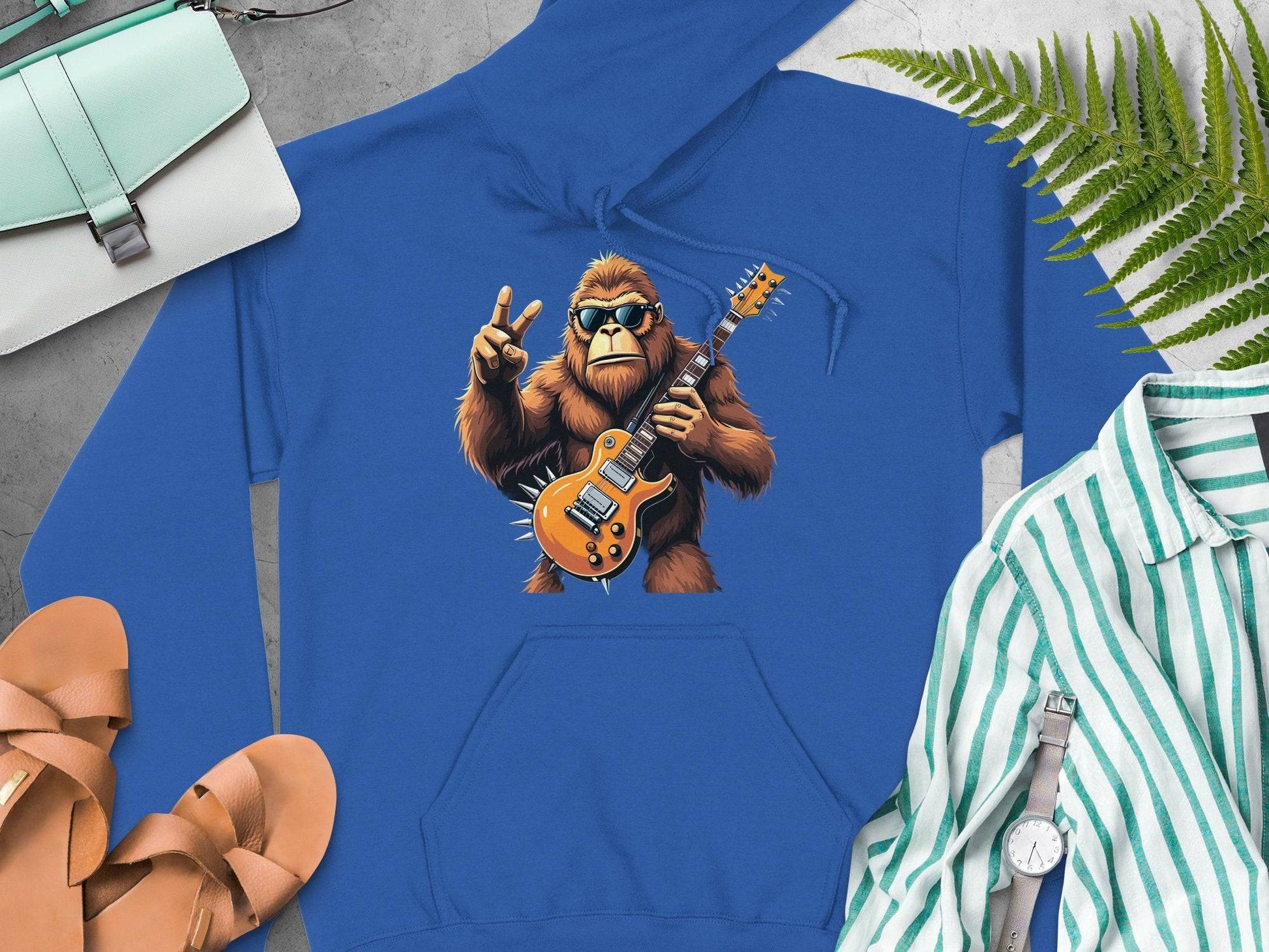 The Garment Graphics gorilla hoodie showcases a graphic of a gorilla playing an electric guitar and flashing a peace sign, ideal for music lovers. Style it with accessories like a teal bag, tan sandals, striped shirt, watch, and green fern decorations.