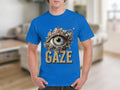 A person is wearing a Garment Graphics classic-fit blue t-shirt with an ornate eye illustration at the center. The word GAZE is boldly printed below the eye, set against a blurred indoor background that emphasizes the unique design.