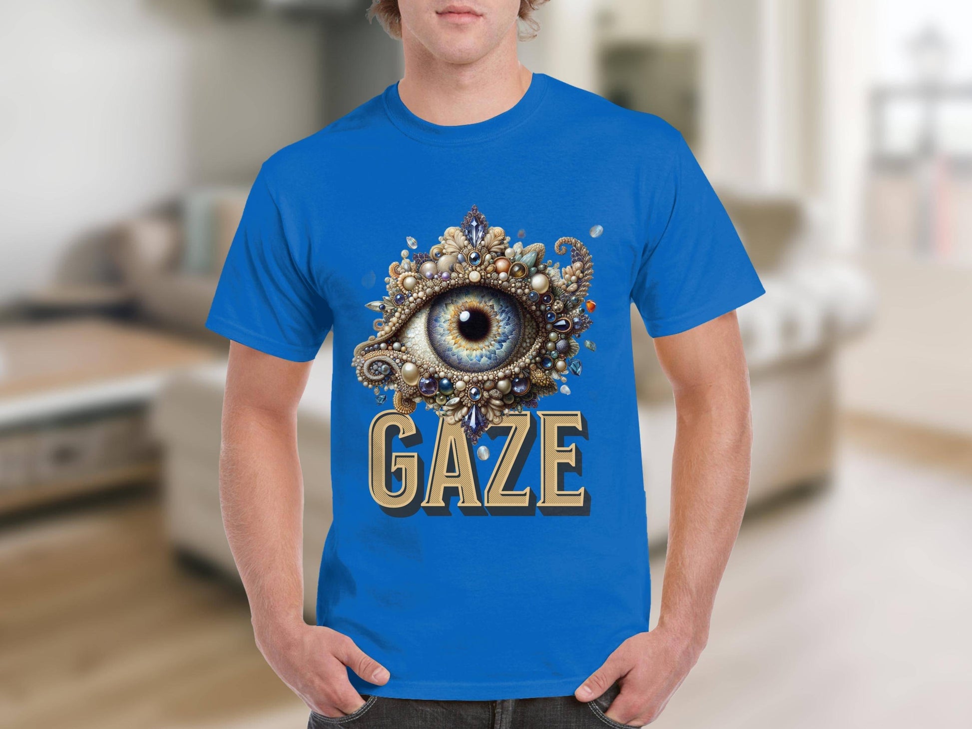 A person is wearing a Garment Graphics classic-fit blue t-shirt with an ornate eye illustration at the center. The word GAZE is boldly printed below the eye, set against a blurred indoor background that emphasizes the unique design.