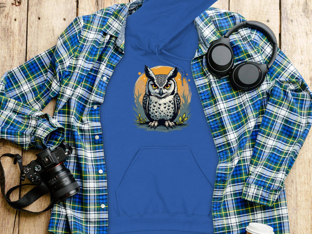 A Garment Graphics classic fit hoodie featuring an owl graphic is set on a wooden surface next to a green and blue plaid shirt, black headphones, a camera, and a white to-go cup. The medium-heavy fabric ensures comfort and style.