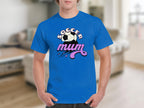 A person wears a classic fit blue Garment Graphics t-shirt reading Soccer mum, featuring a graphic design with a soccer ball and stars. The background is a cozy living room with a sofa and blurred elements.