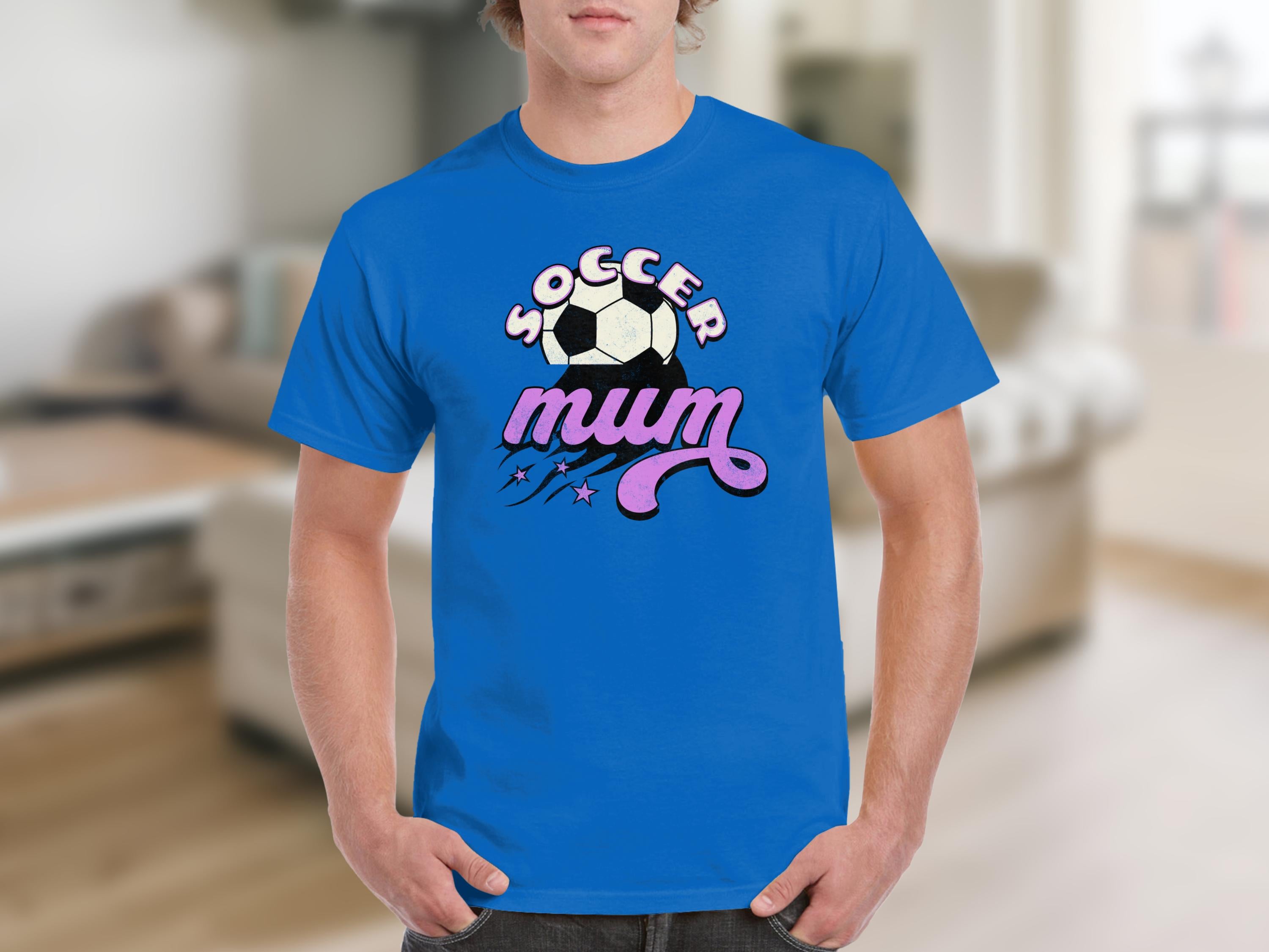 A person wears a classic fit blue Garment Graphics t-shirt reading Soccer mum, featuring a graphic design with a soccer ball and stars. The background is a cozy living room with a sofa and blurred elements.